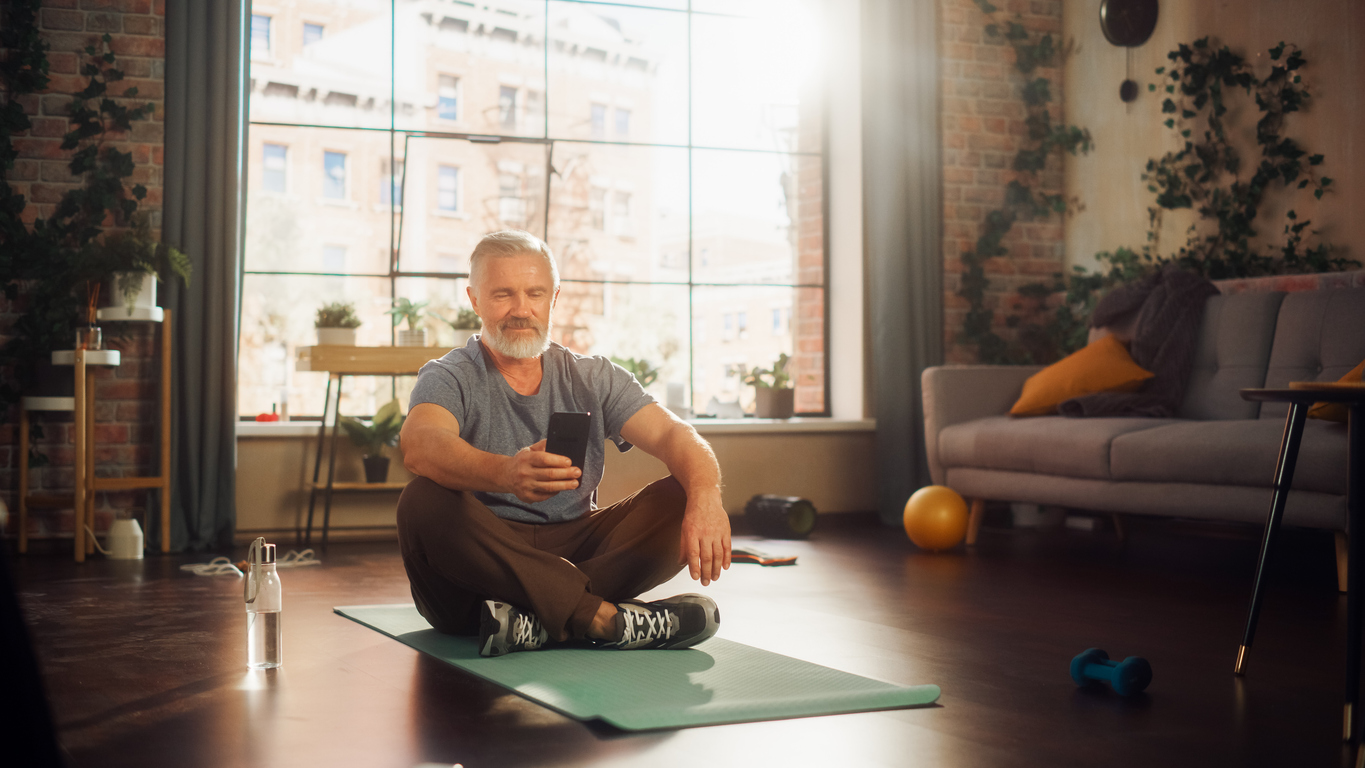 Mann sitzt auf dem Boden auf einer Yogamatte mit einem Smartphone in der Hand. Er lächelt.