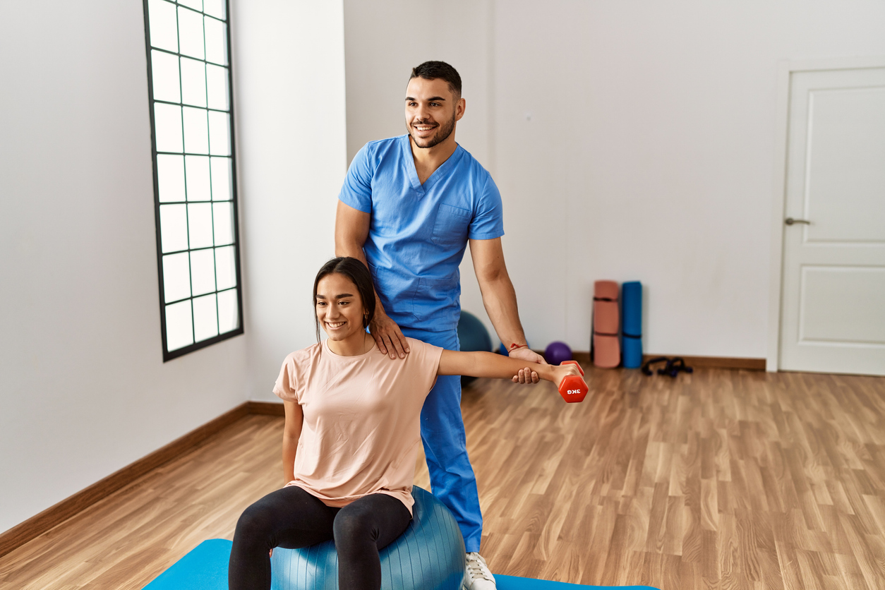 Frau sitzt auf einem Sitzball. Ein Physiotherapeut steht hinter ihr und streckt ihren linken Arm aus. Beide lachen.