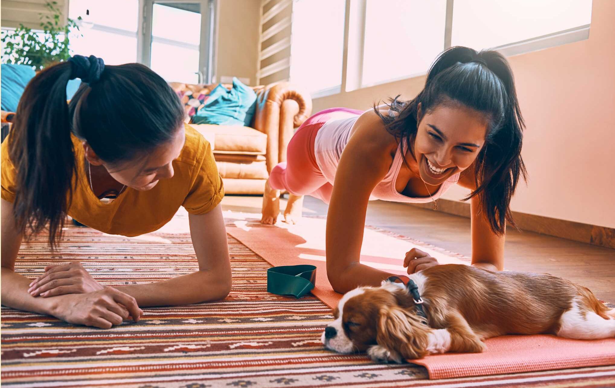 Zwei Frauen liegen auf dem Boden und machen eine Rückenübung. Vor ihnen liegt ein Hund und sie lachen.