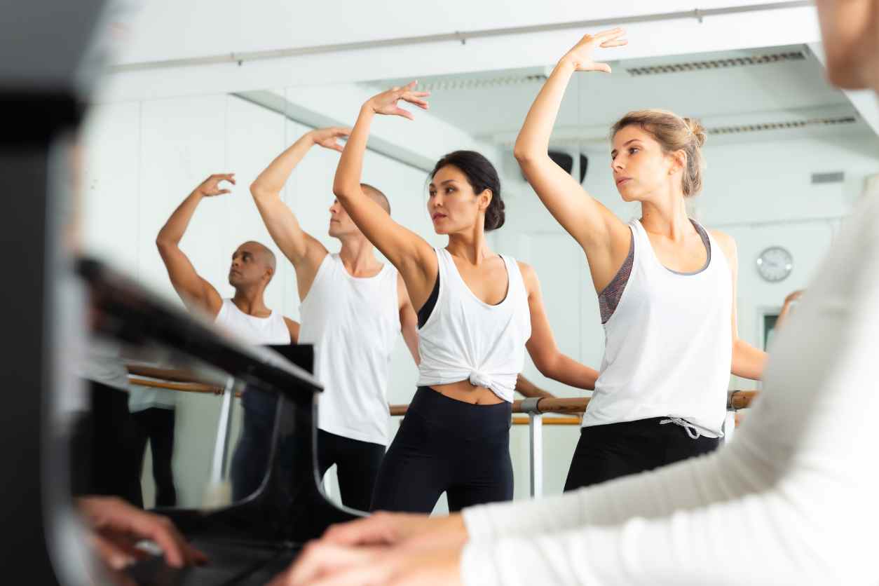 Ballettschüler tanzen an einer Stange mit gehobenen Armen. Sie tragen weiße T-Shirts und schwarze Leggins.