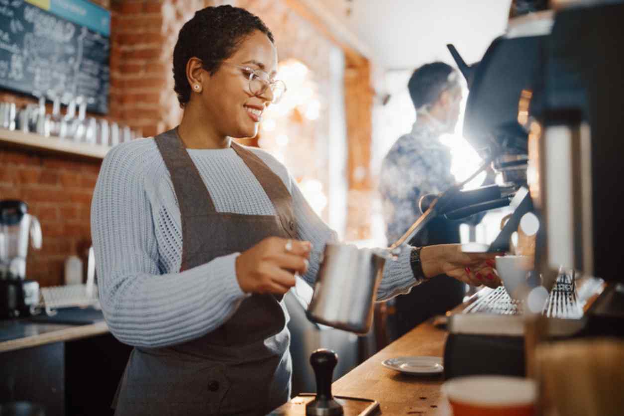 Eine Frau mit dunkler Hautfarbe steht an einer Kaffeemaschine. Sie macht einen Espresso und trägt eine Schürze.