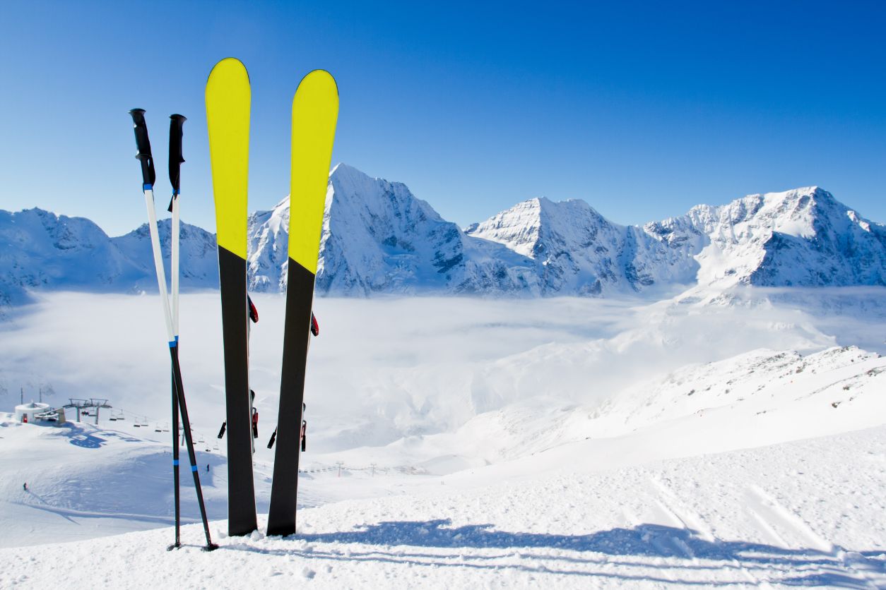 Zwei Ski stecken im Schnee. Sie haben gelbe Spitzen. Im Hintergrund ist eine verschneite Bergkette zu sehen und ein Skilift.