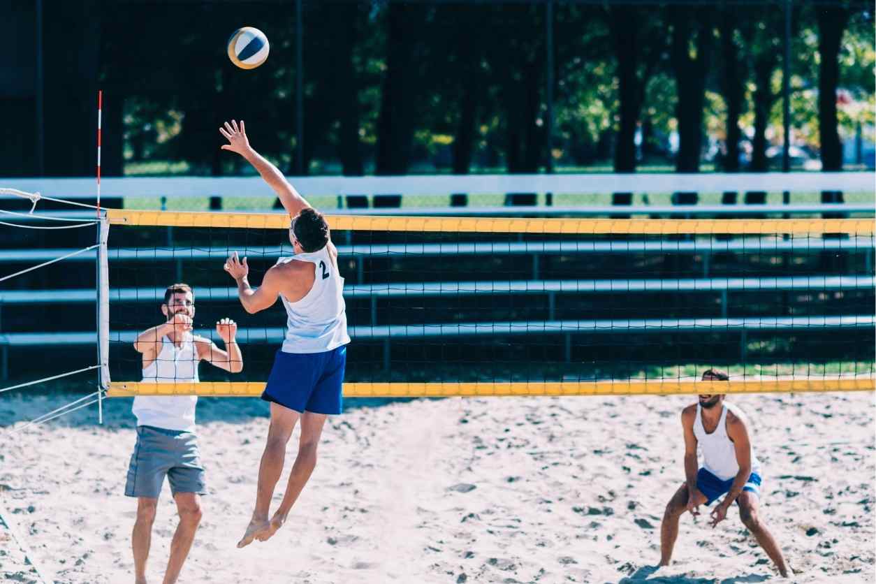 Drei Männer spielen auf einem Beachvolleyballfeld. Einer springt gerade in die Luft, man sieht ihn von hinten. Der Ball fliegt gerade übers Netz.